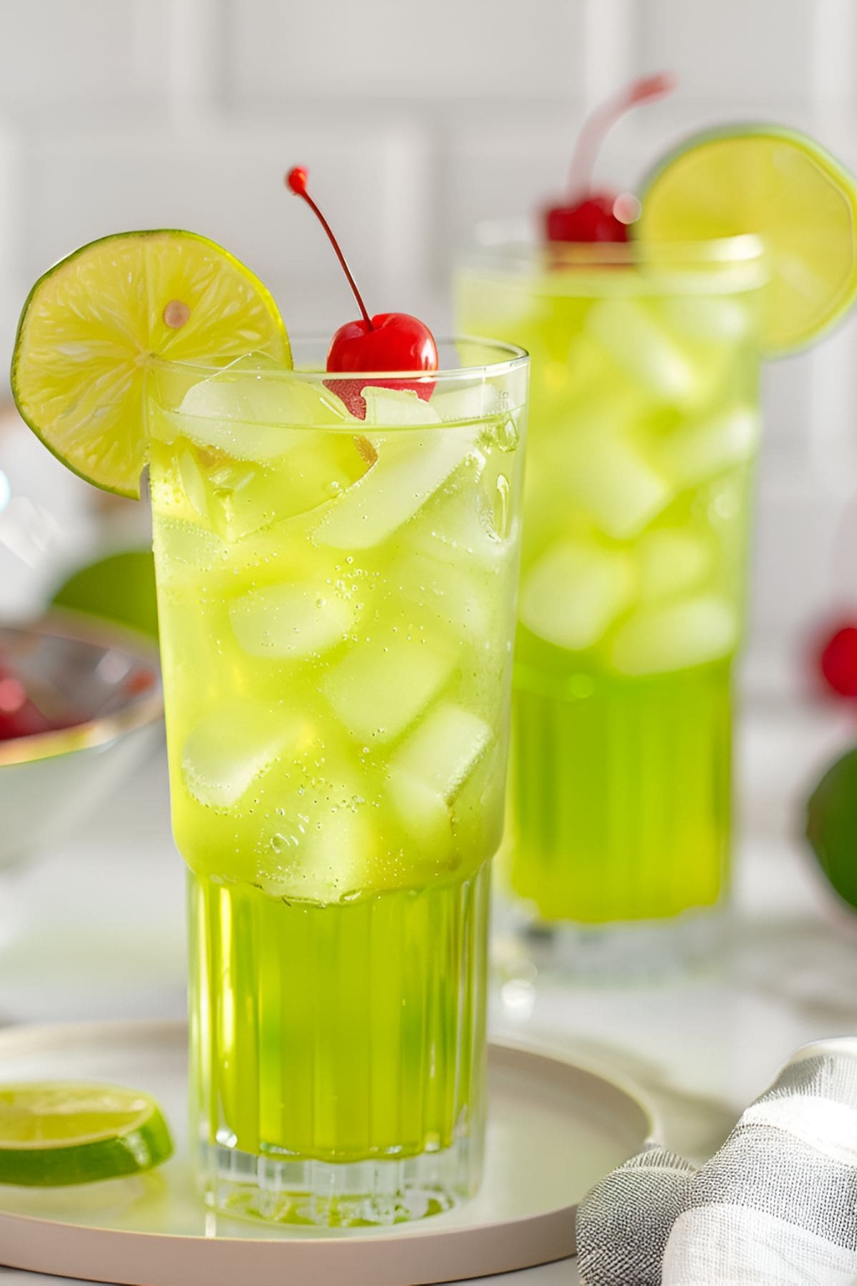 Two Glasses of Bright Green Tokyo Tea Cocktails in Tall Glasses with Ice, Cherries for Garnish, and a Lime Wheel