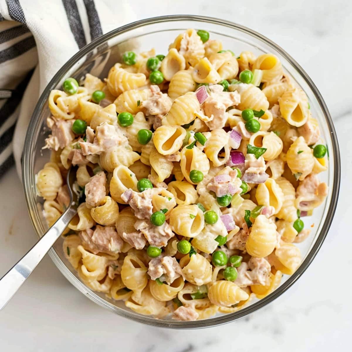 Homemade salad featuring pasta shells, tuna, green peas, red onions in a glass bowl.