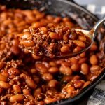 Close Up of a Spoonful of BBQ Baked Beans- with Ground Beef, Beans, and Sauce- Held Over a Cast Iron Skillet with More BBQ Baked Beans