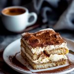 Close up of a Slice of Tiramisu with Layers of Ladyfinger Cookies, Mascarpone Whipped Cream, and Cocoa Powder Dusting on a Plate with a Mug of Coffee in the Background