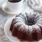 Too Much Chocolate Cake Bundt on a Cake Plate, Dusted with Powdered Sugar, with a Mug of Coffee in the Background