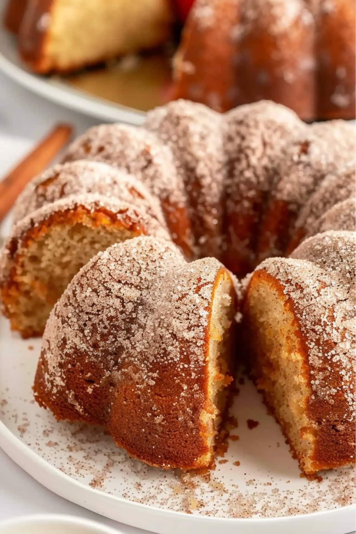 Sliced whole apple cider donut bundt cake on white plate sprinkled with cinnamon sugar.