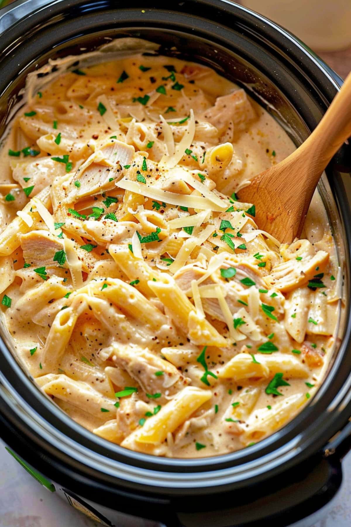 Crockpot with penne pasta cooked in creamy sauce and shredded chicken garnished with shredded parmesan cheese.