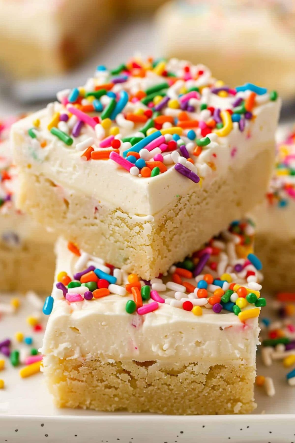 Square slices of frosted cookie bars with candy sprinkles stacked on a white plate.