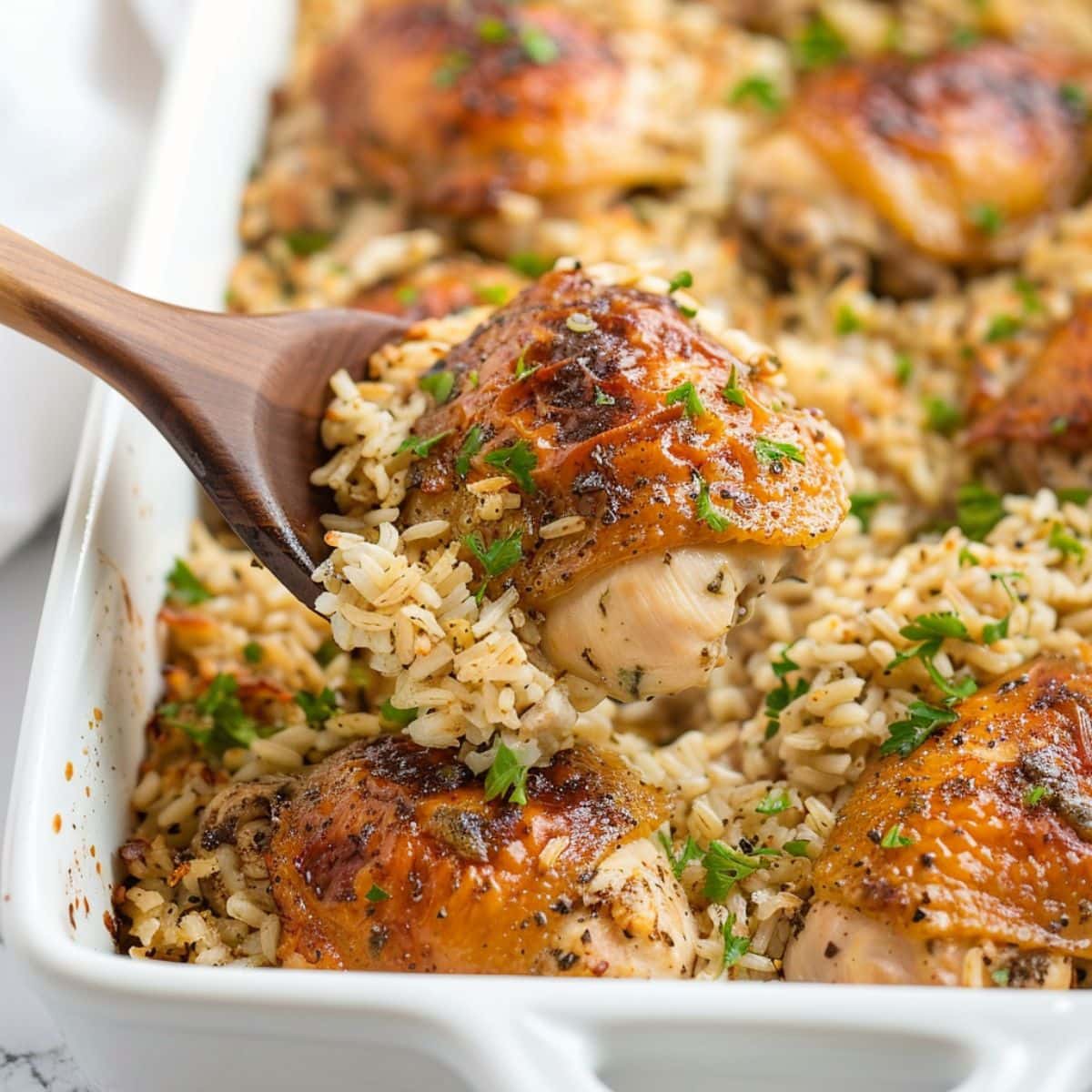 Wooden ladle lifting a serving of oven  baked chicken and rice on a white baking dish.