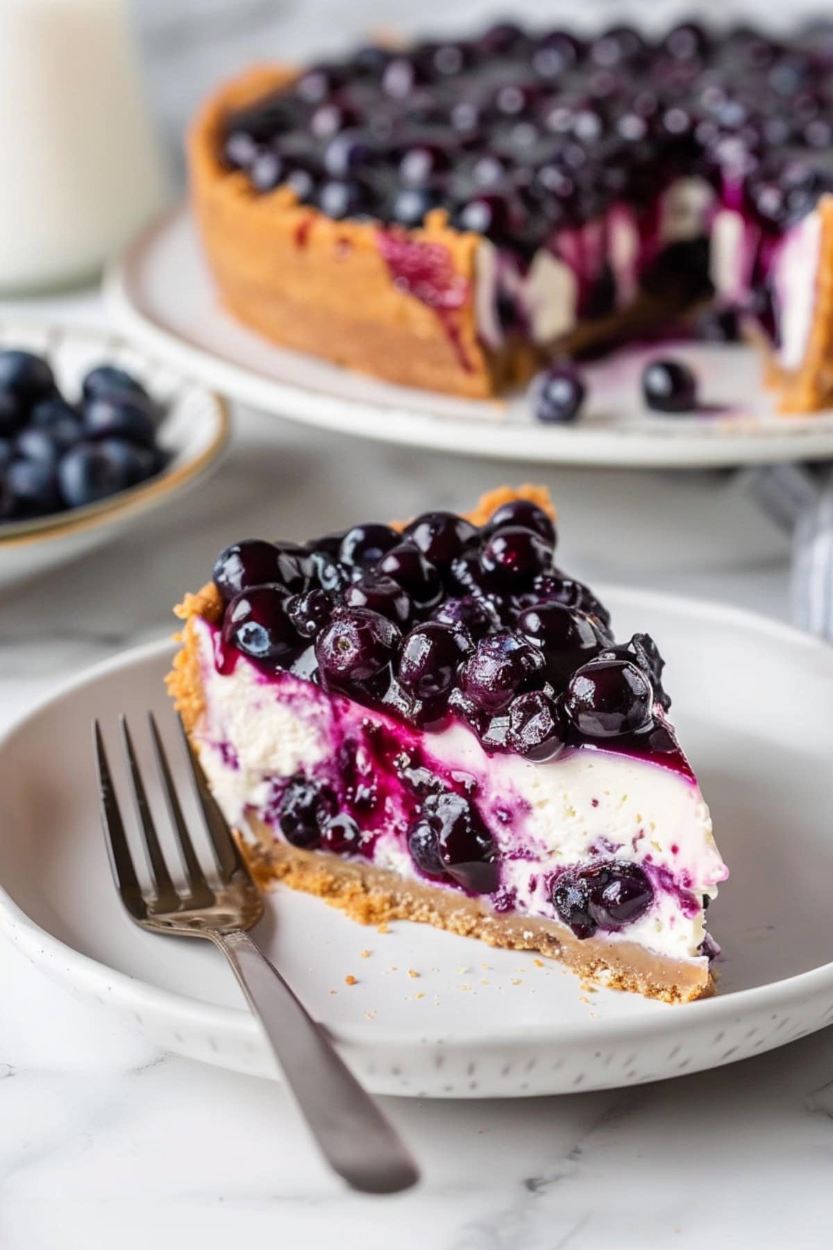 Sliced lemon blueberry cheesecake on a white plate, with a whole cheesecake in the background.