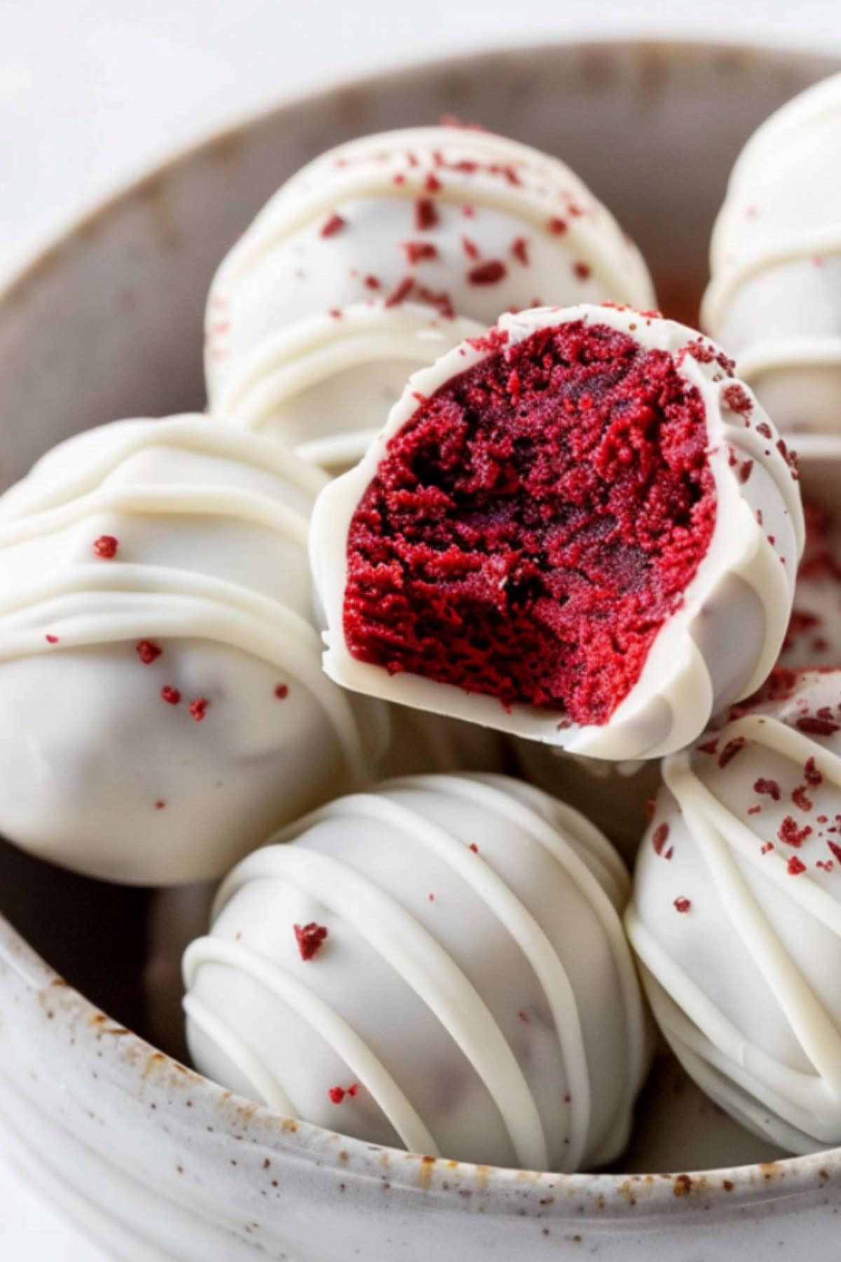 A red velvet truffle, cut in half, showing its soft, creamy red filling and white chocolate exterior. It's in a bowl of red velvet truffles
