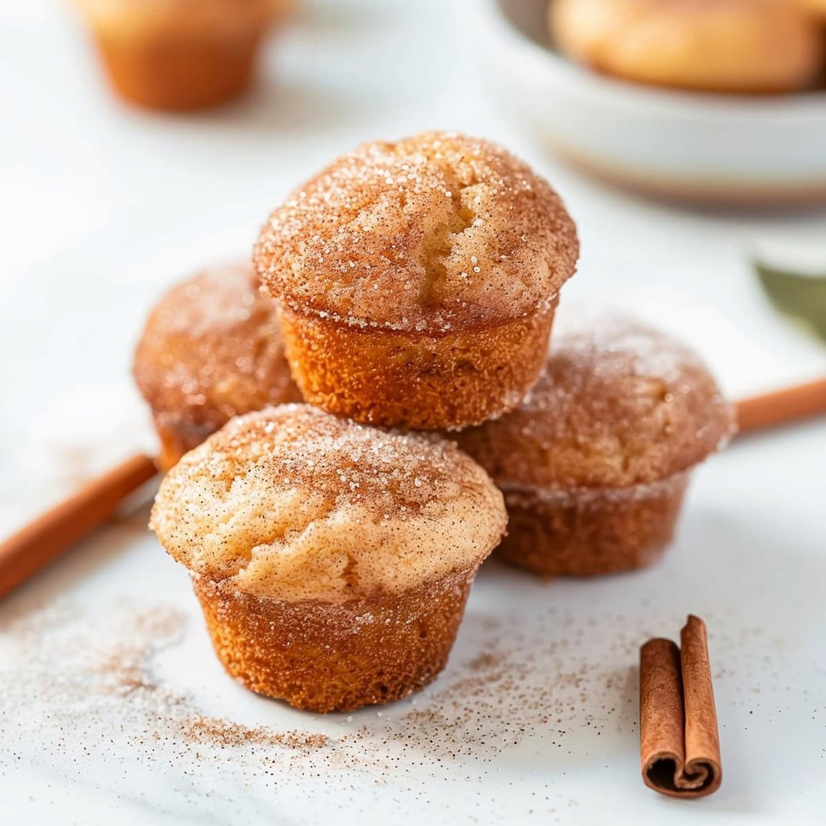 Snickerdoodle muffins coated with cinnamon sugar stacked on a white marble table with cinnamon sticks on the side 