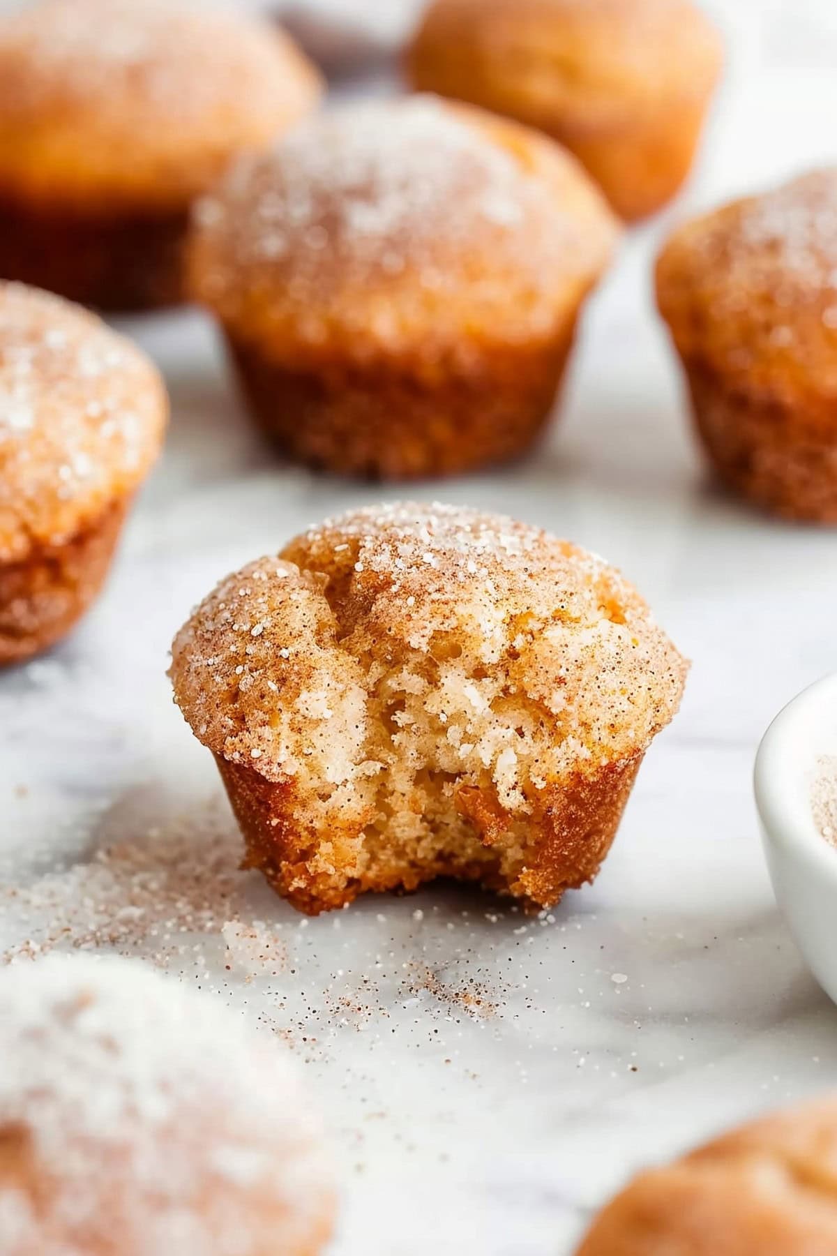 Snickerdoodle muffins sitting on a white marble surface with a bite taken out of one.