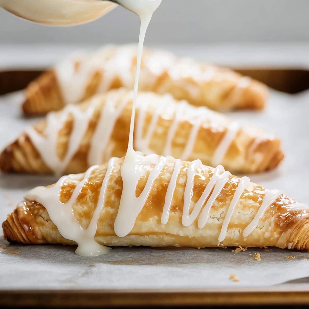 Warm apple turnovers with a buttery, flaky pastry on a baking sheet being drizzled with sweet vanilla glaze, side view