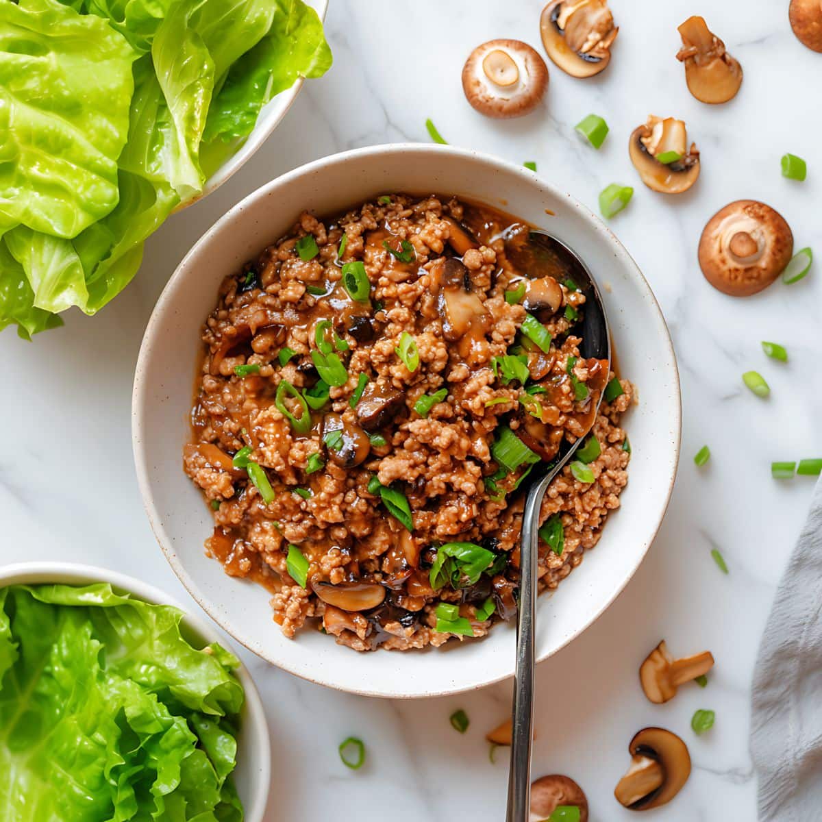 Sauteed ground chicken in a white bowl, fresh lettuce and mushrooms flat lay on a white marble table. 