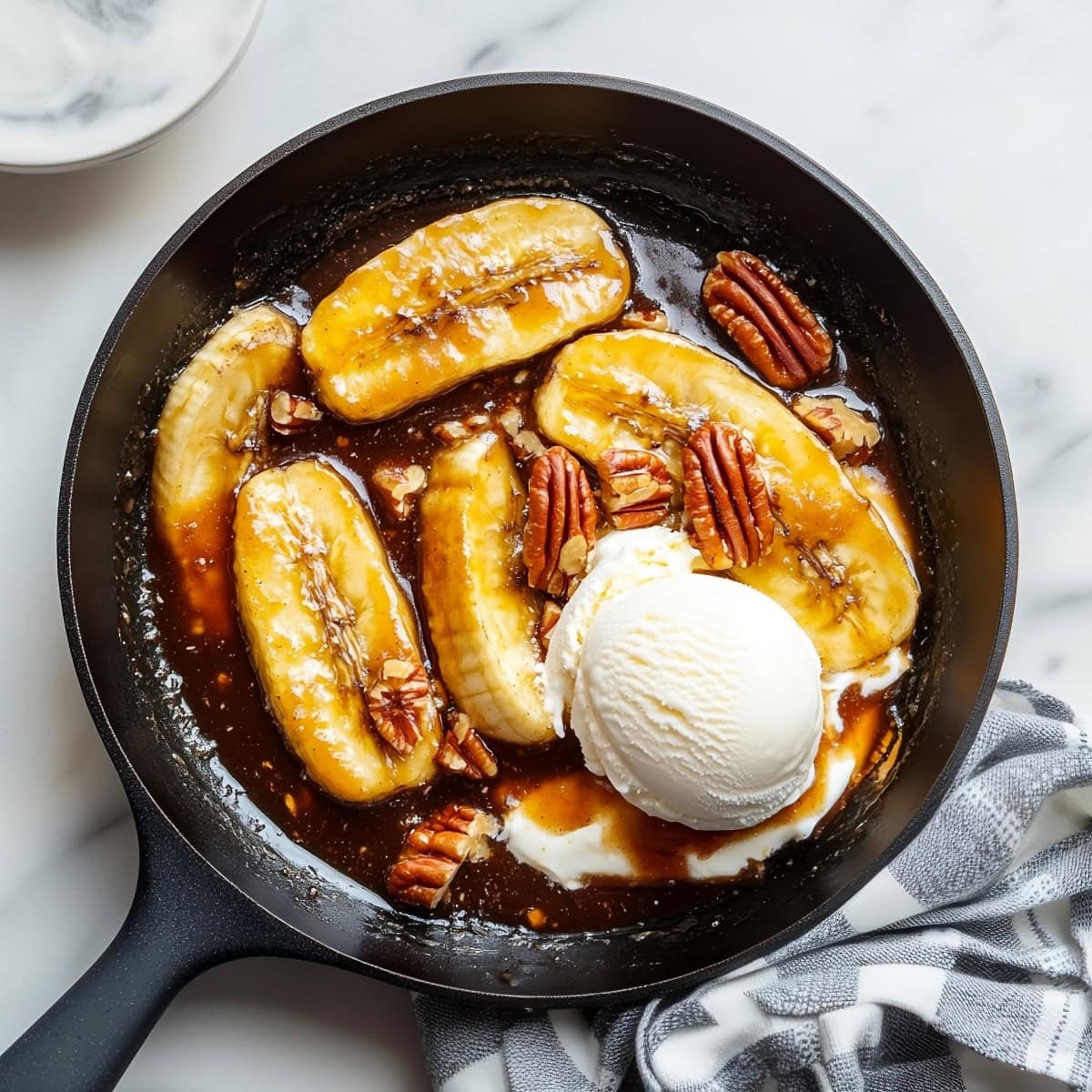 Overhead view of caramelized bananas foster in a black skillet with ice cream and pecans.