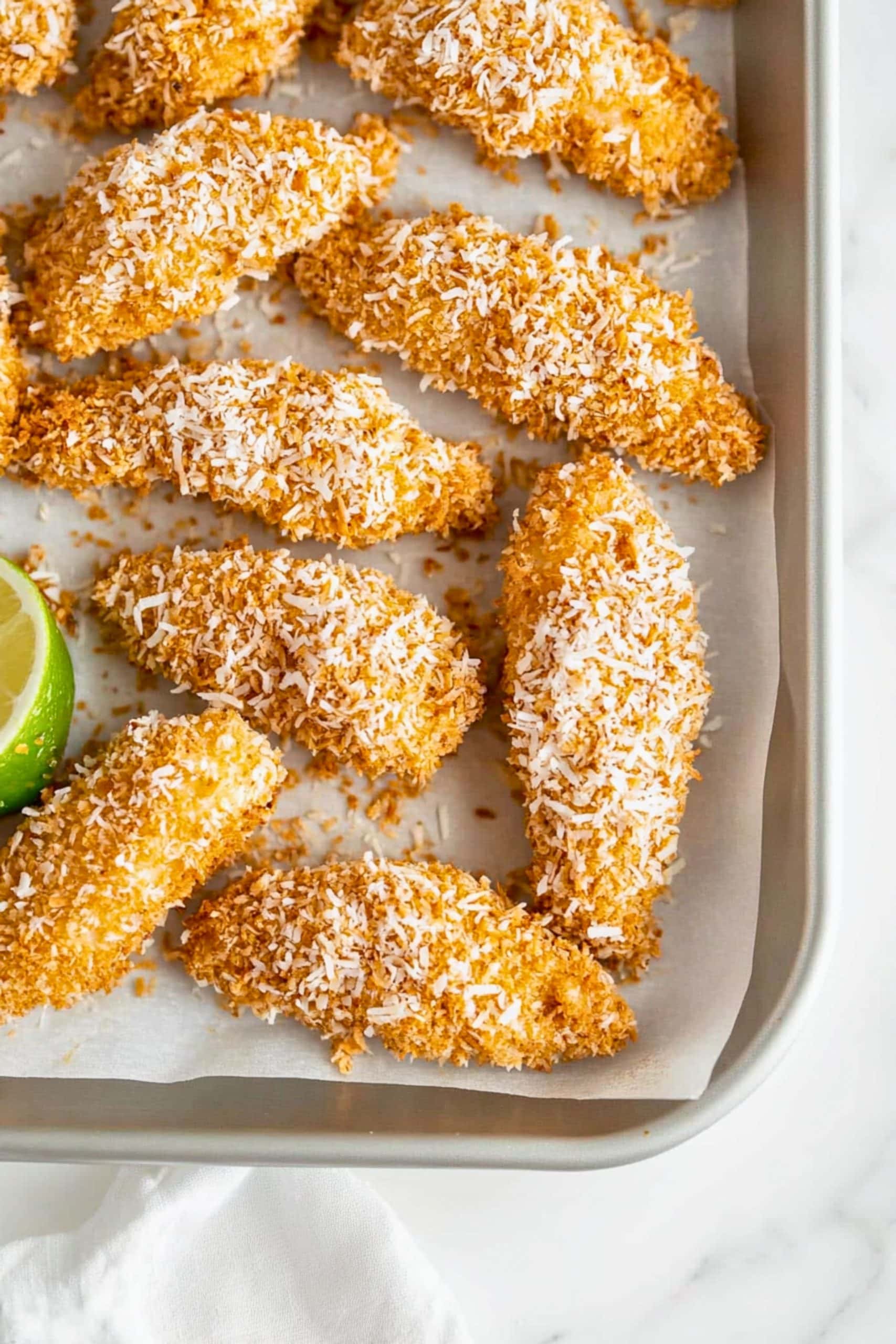 Coconut and bread crumbs coated chicken tenders arranged in a bakign sheet.