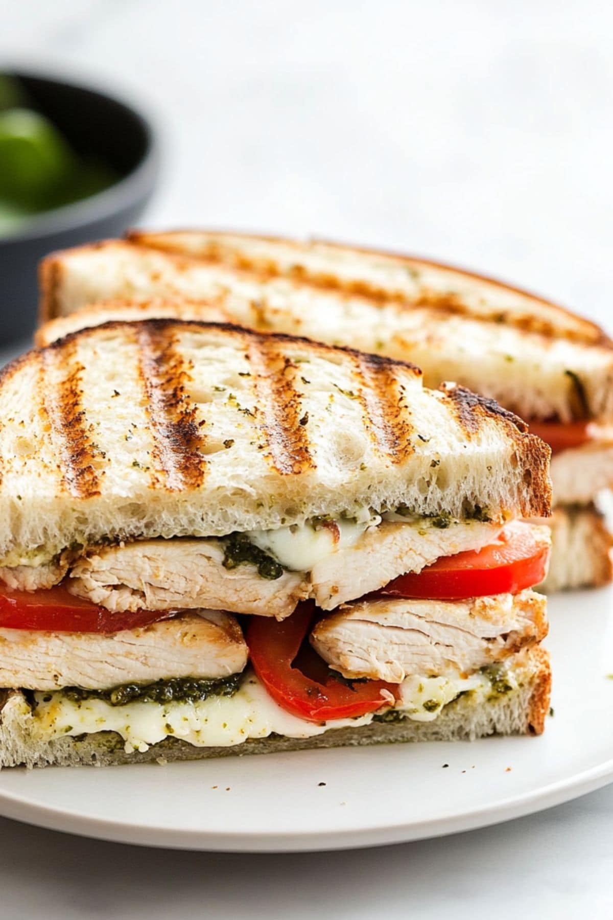 grilled chicken panini cut in half on a plate, with juicy chicken, sliced tomatoes, pesto, and mozzarella cheese. 