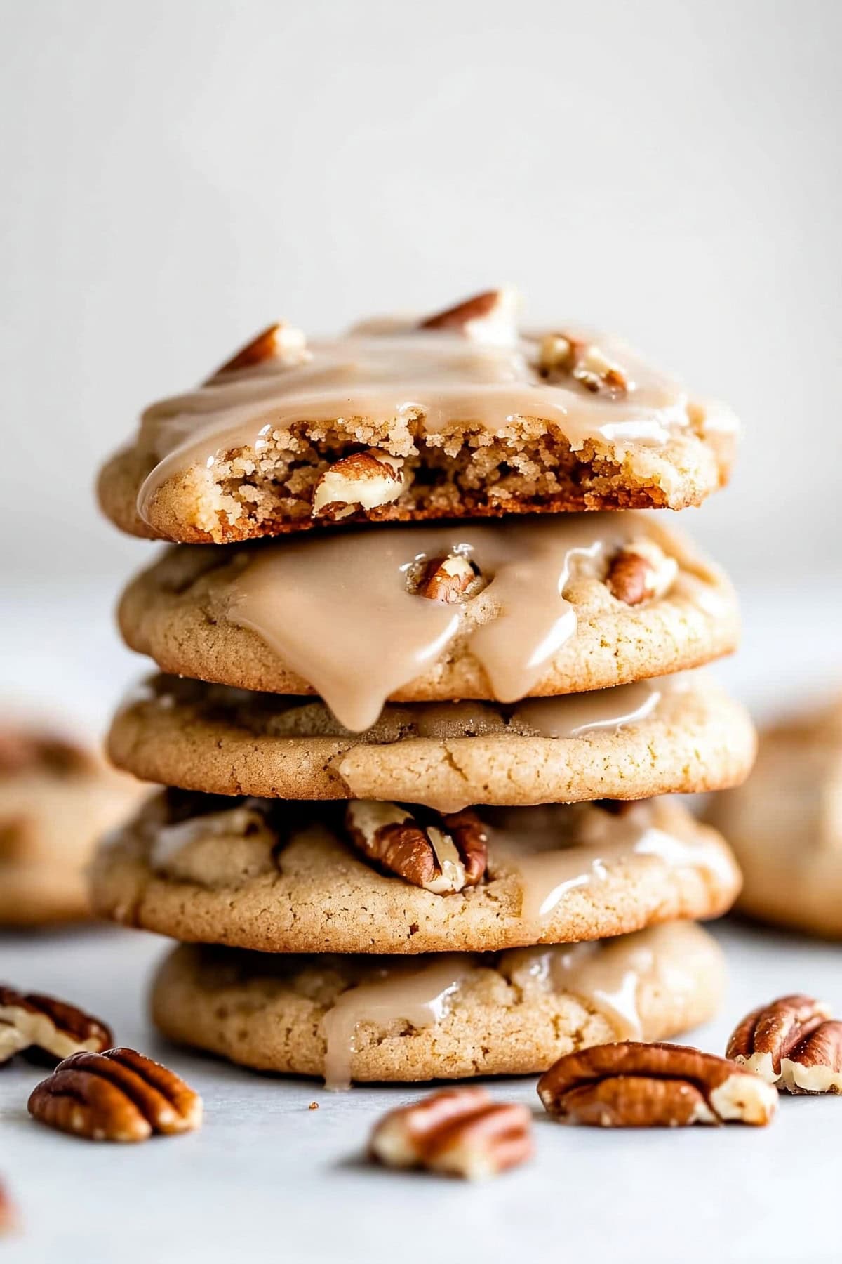 Maple cookies with maple icing and chopped pecans stacked on a white marble table.