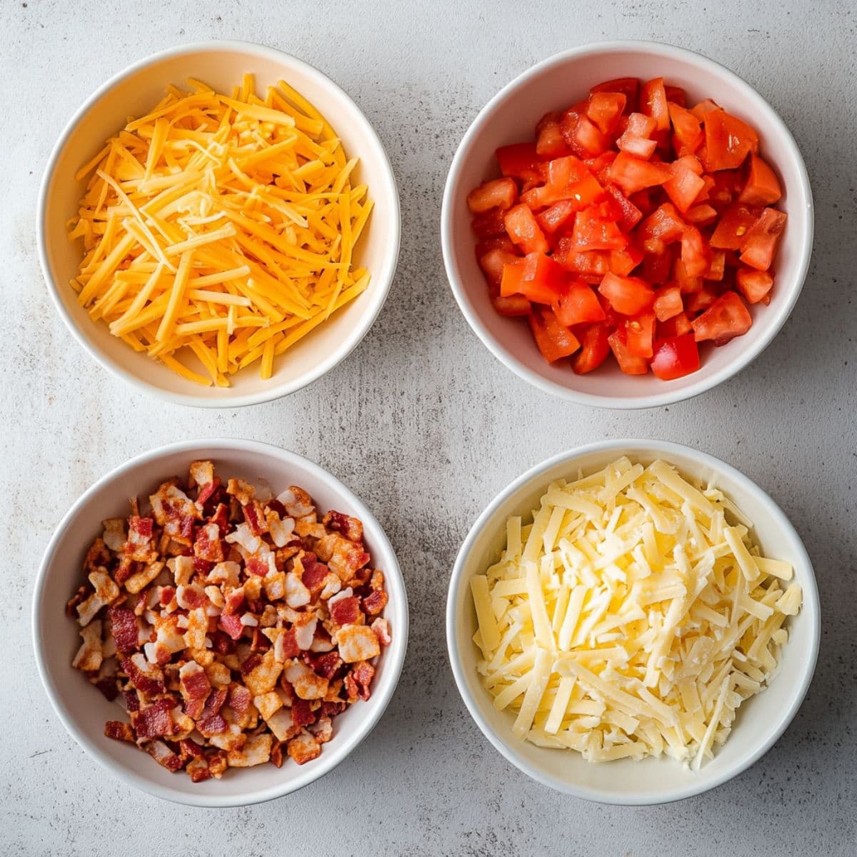 Four bowls with shredded cheese, diced tomato and crumbled bacon on a concrete table, top down view.