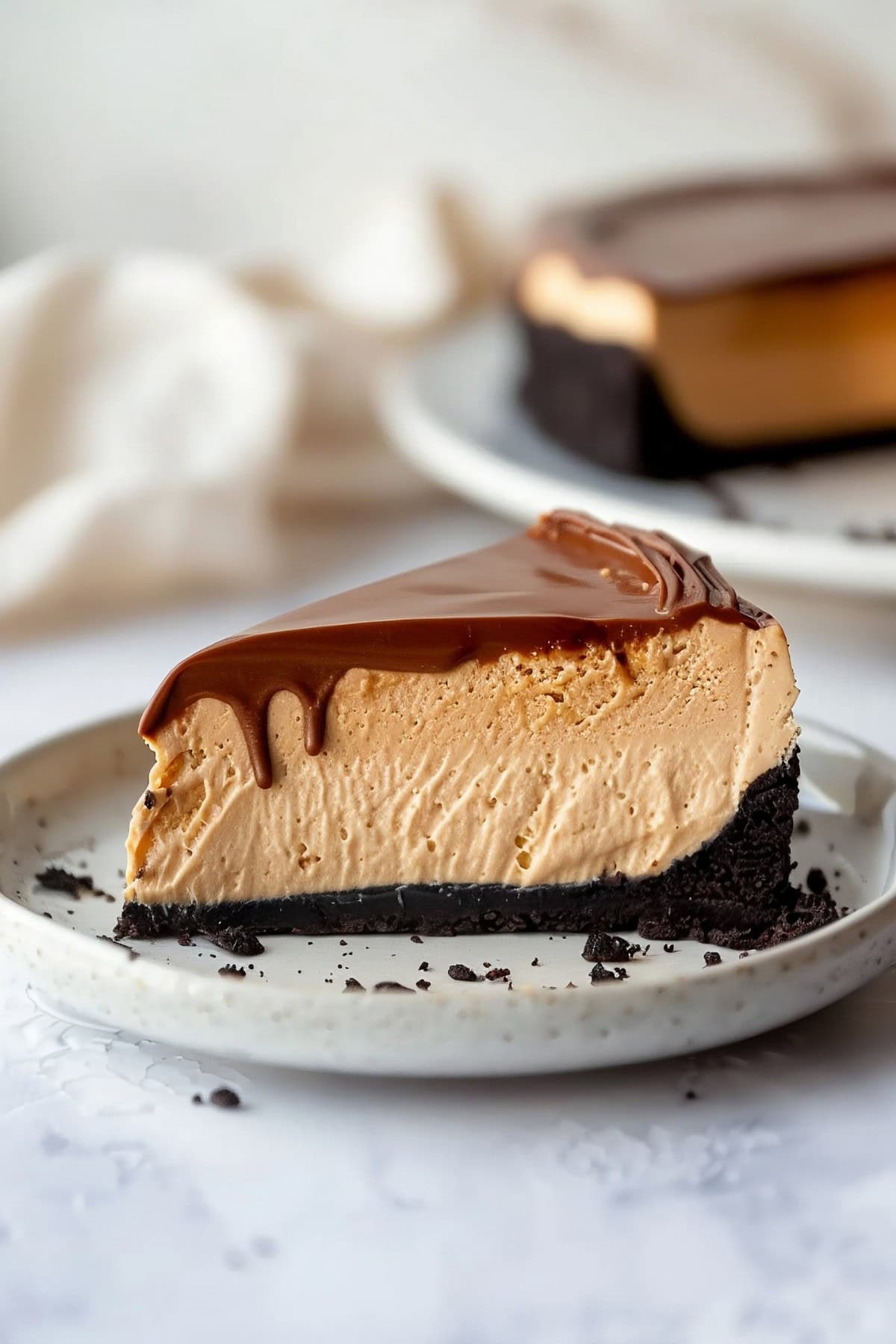 Slice of homemade peanut butter cheesecake with Oreo crust and ganache topping on a plate, side view