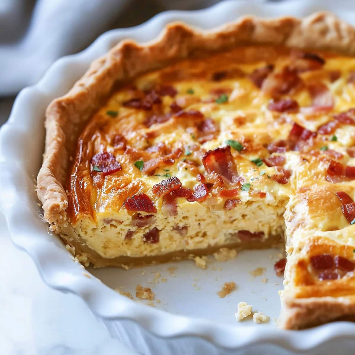 Classic quiche Lorraine with a golden, flaky crust and a savory filling in a white pie dish and a slice removed.