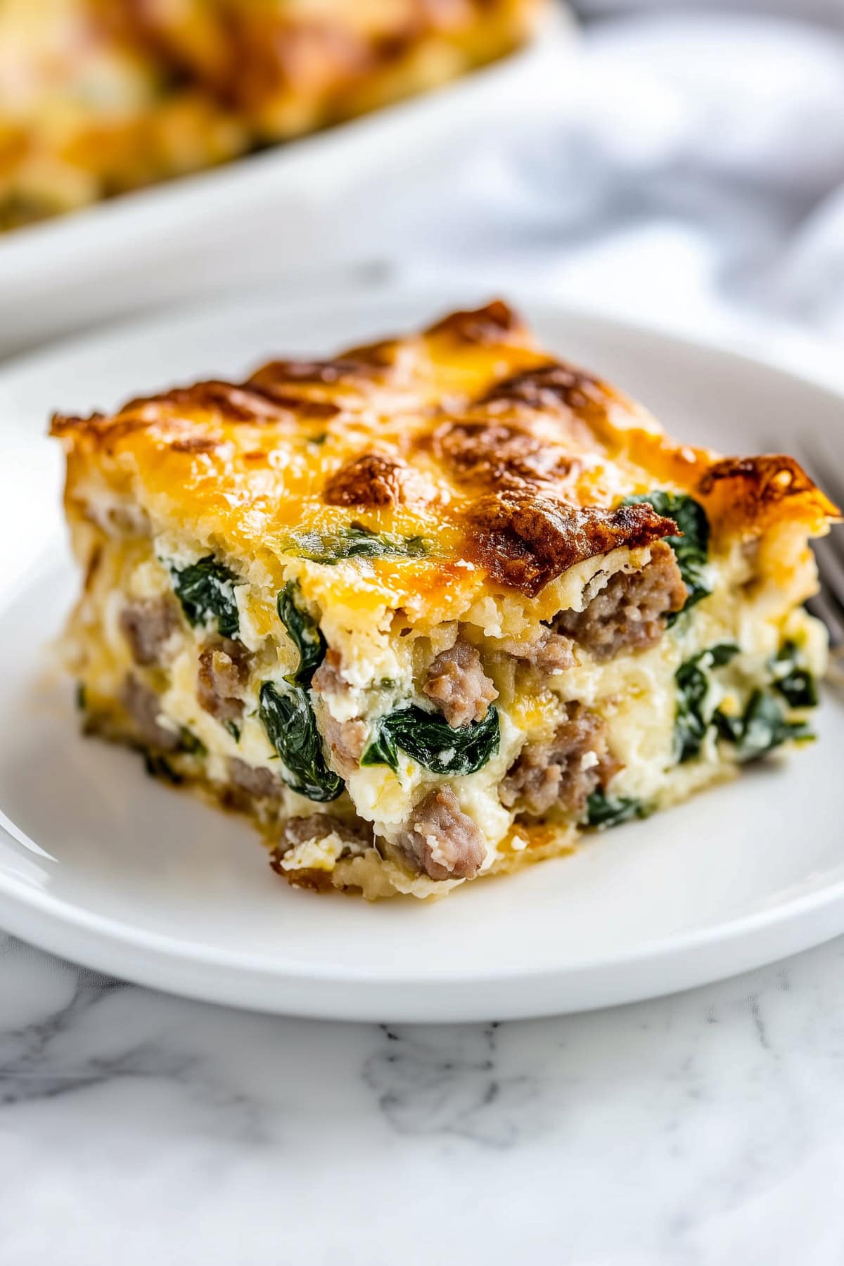 A piece of breakfast strata with spinach and sausage in a white plate on a white marble table.