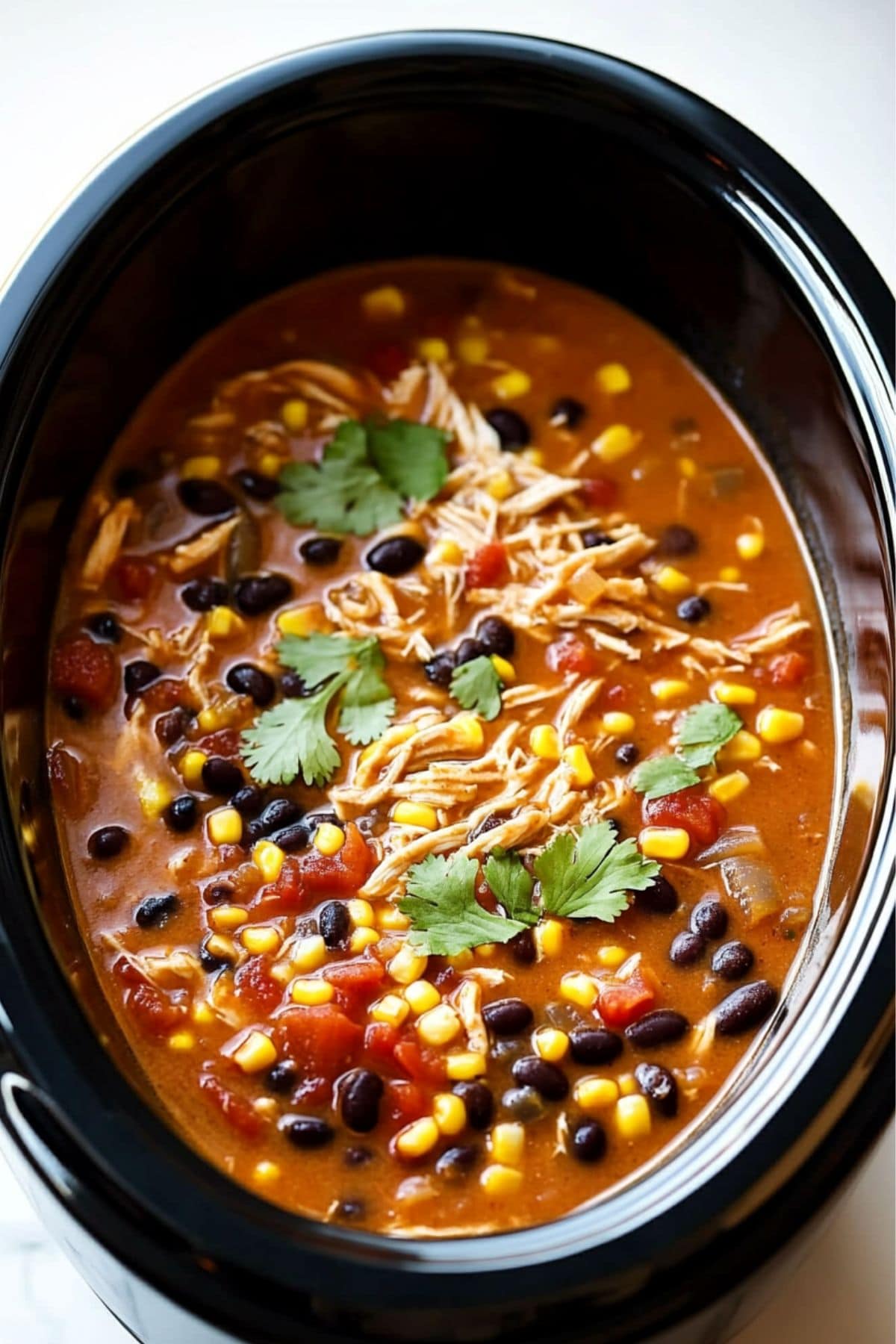 Shredded chicken, corn and black beans, lifting a serving of chicken enchilada soup in a slow cooker sitting on a white marble table.