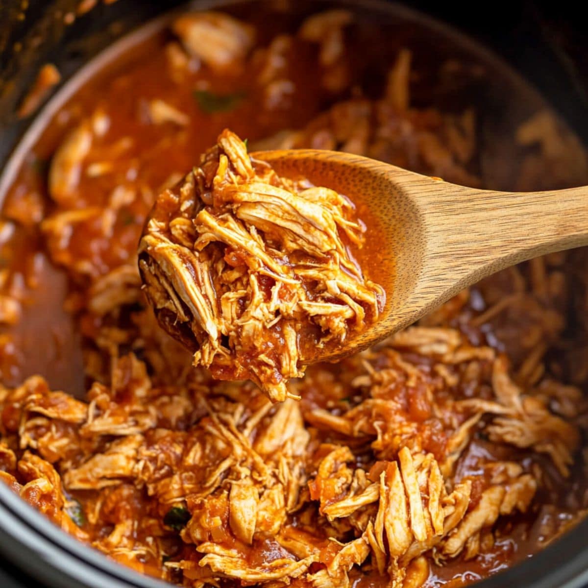 Focus shot of wooden spoon with scoop of chicken tinga cooked in a slow cooker.