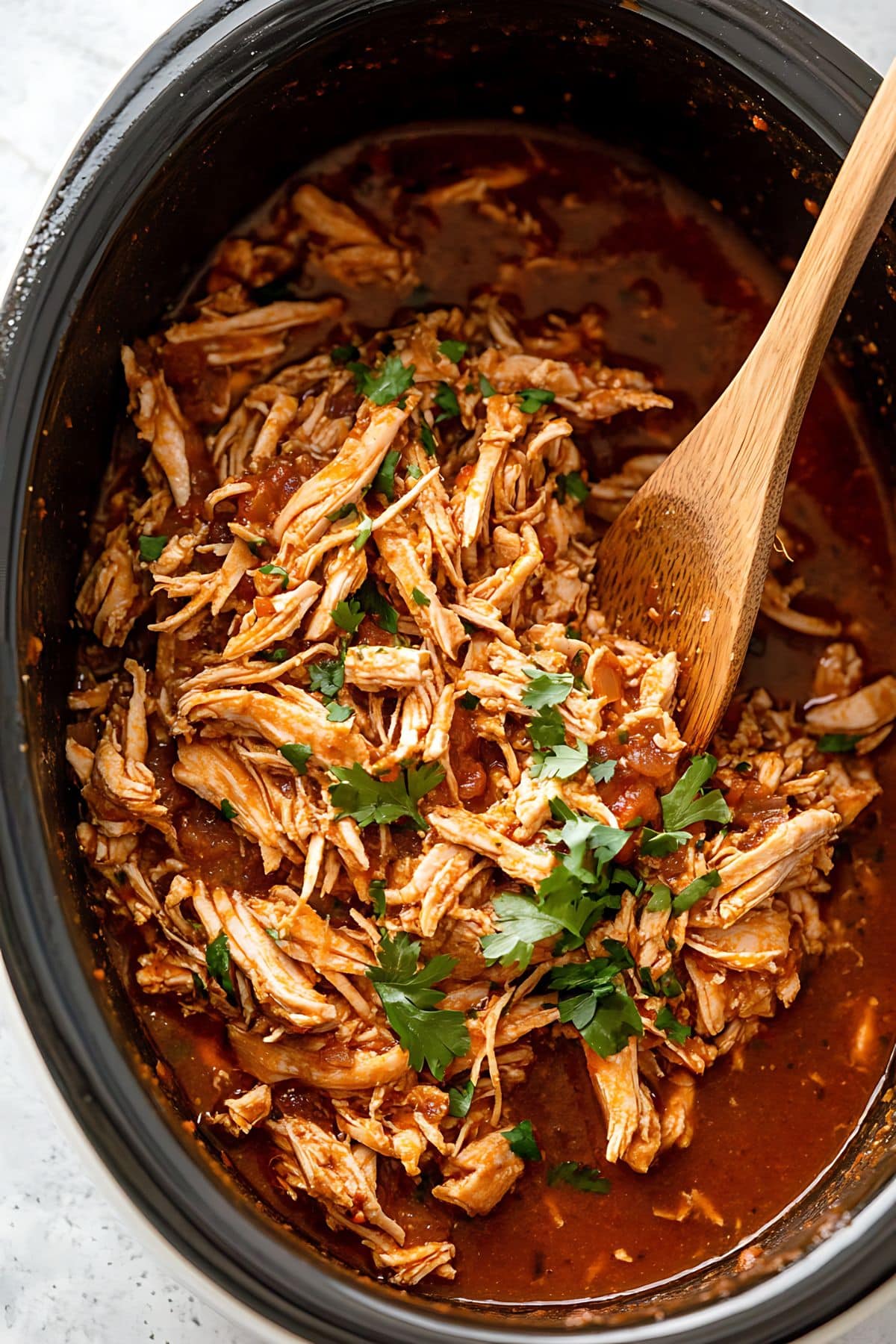 Wooden spoon ladle tossing shredded chicken with tomato sauce in a slow cooker.