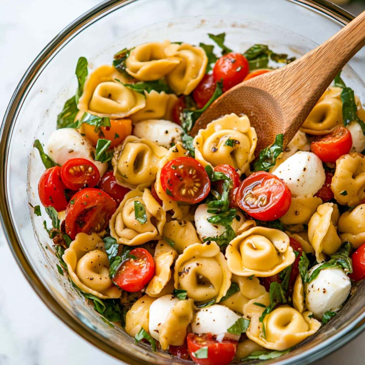 Tortellini caprese salad tossed by a wooden spoon in a large glass mixing bowl.