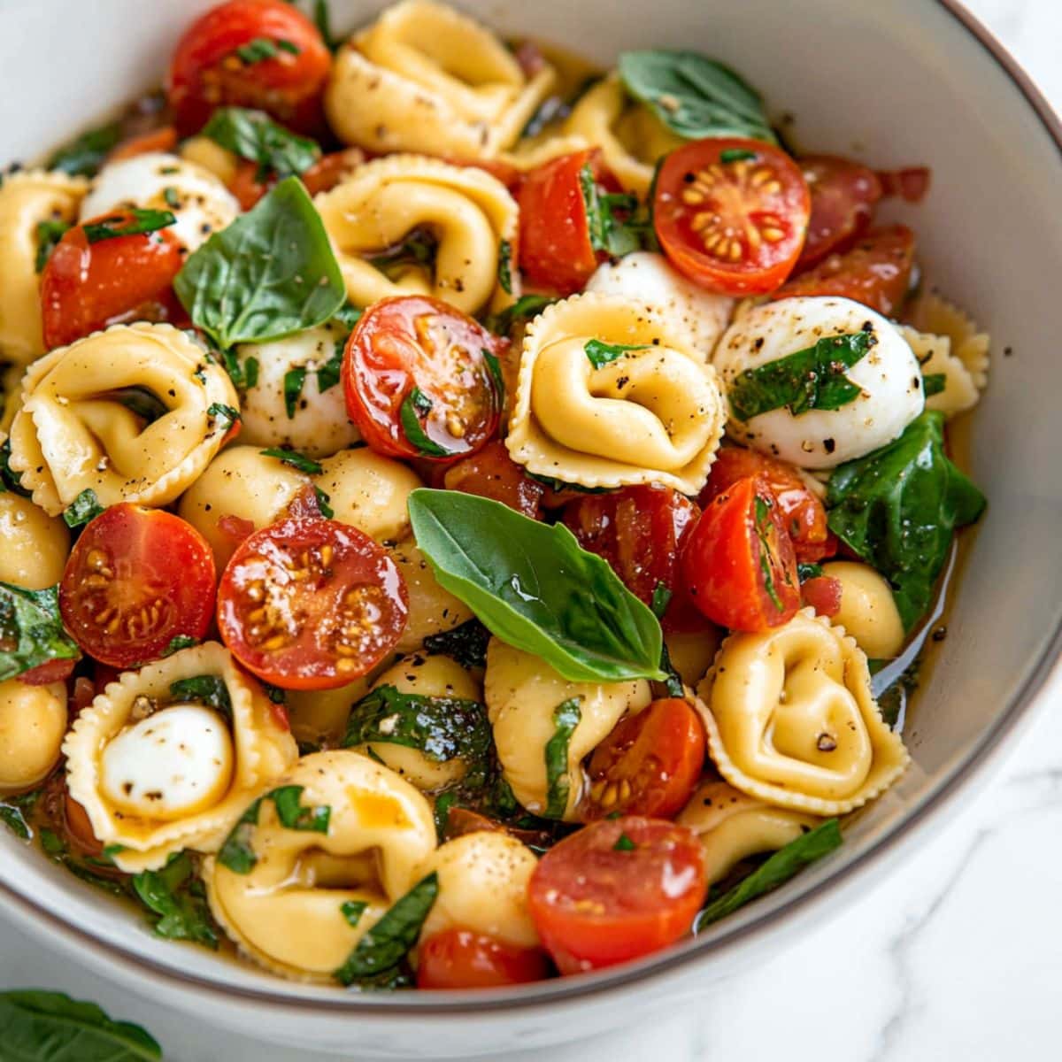 Cheese tortellini with jcherry tomatoes, mozzarella pearls, and chopped fresh basil tossed in balsamic dressing served on a white bowl. 
