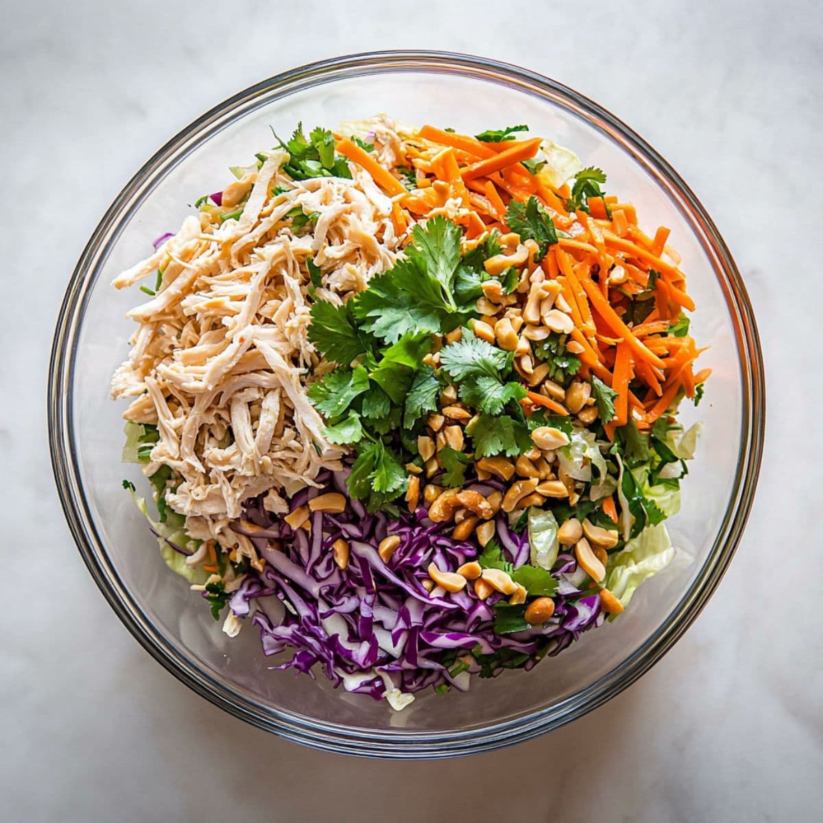 Shredded cabbage, carrots, onions, cilantro leaves and chopped peanuts arranged inside a large glass mixing bowl.