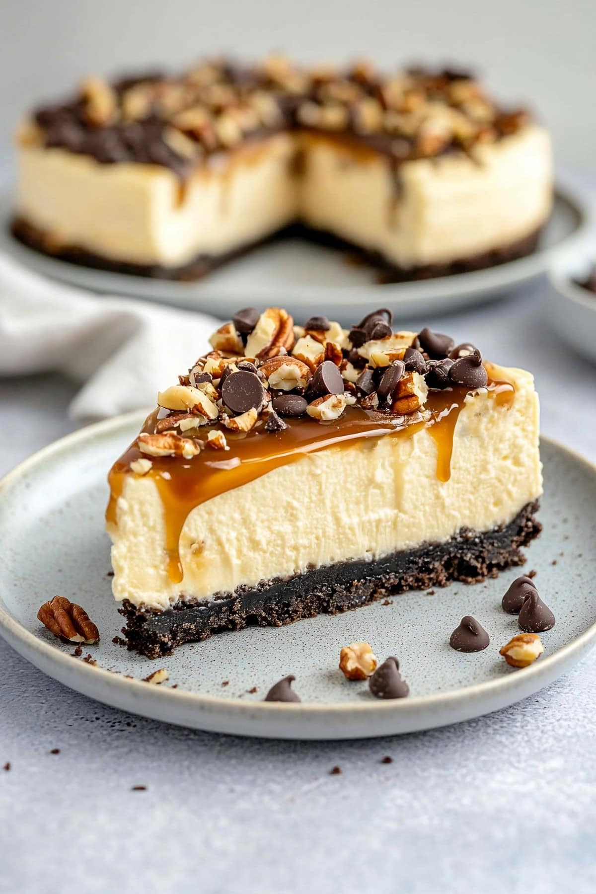 A slice of caramel turtle cheesecake on a white plate garnished with caramel sauce, chopped pecans, and chocolate chips