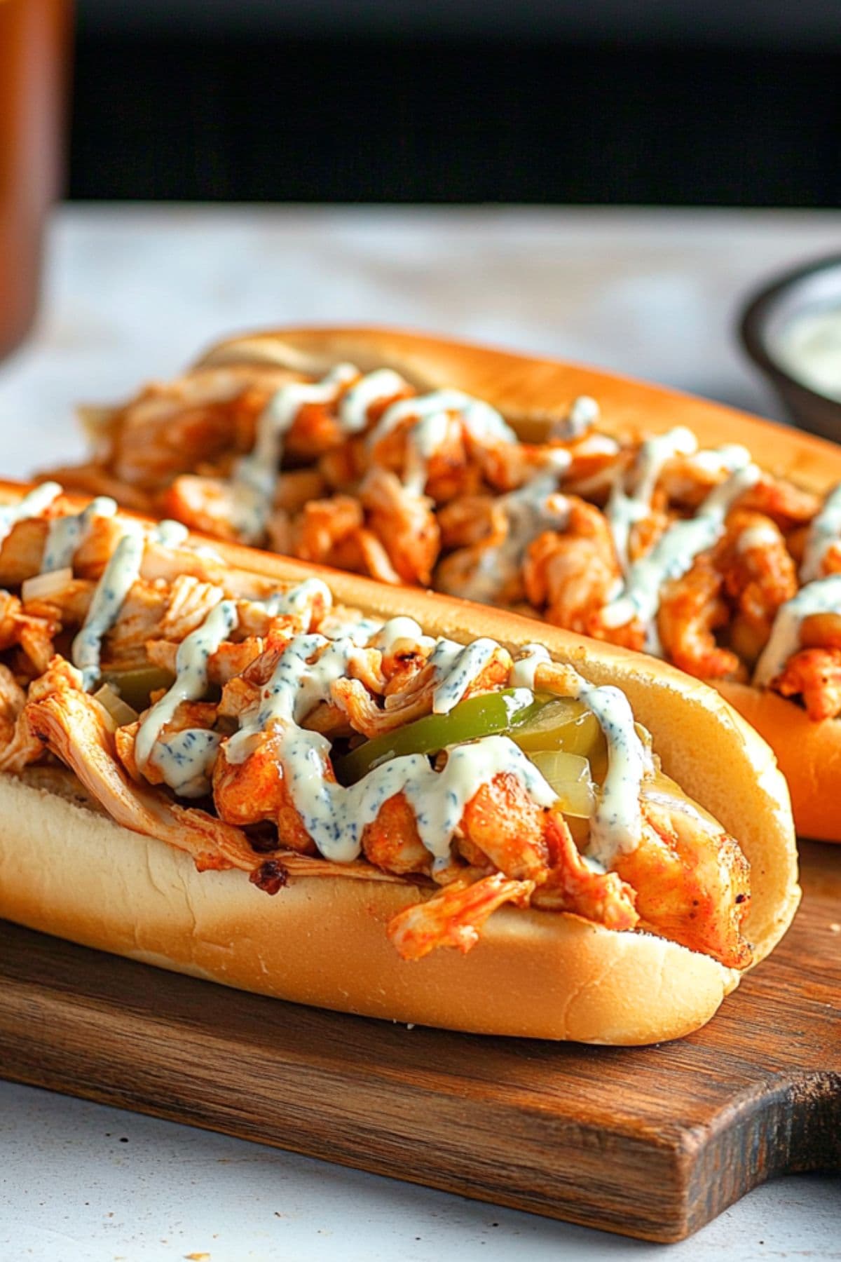 Two Buffalo Chicken Cheesesteaks in hoagie rolls with blue cheese dressing on a chopping board