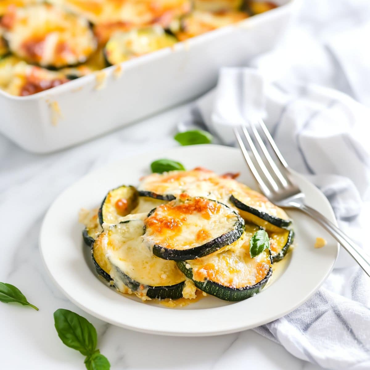 Serving of cheesy scalloped zucchini on a white plate with fork on the side.