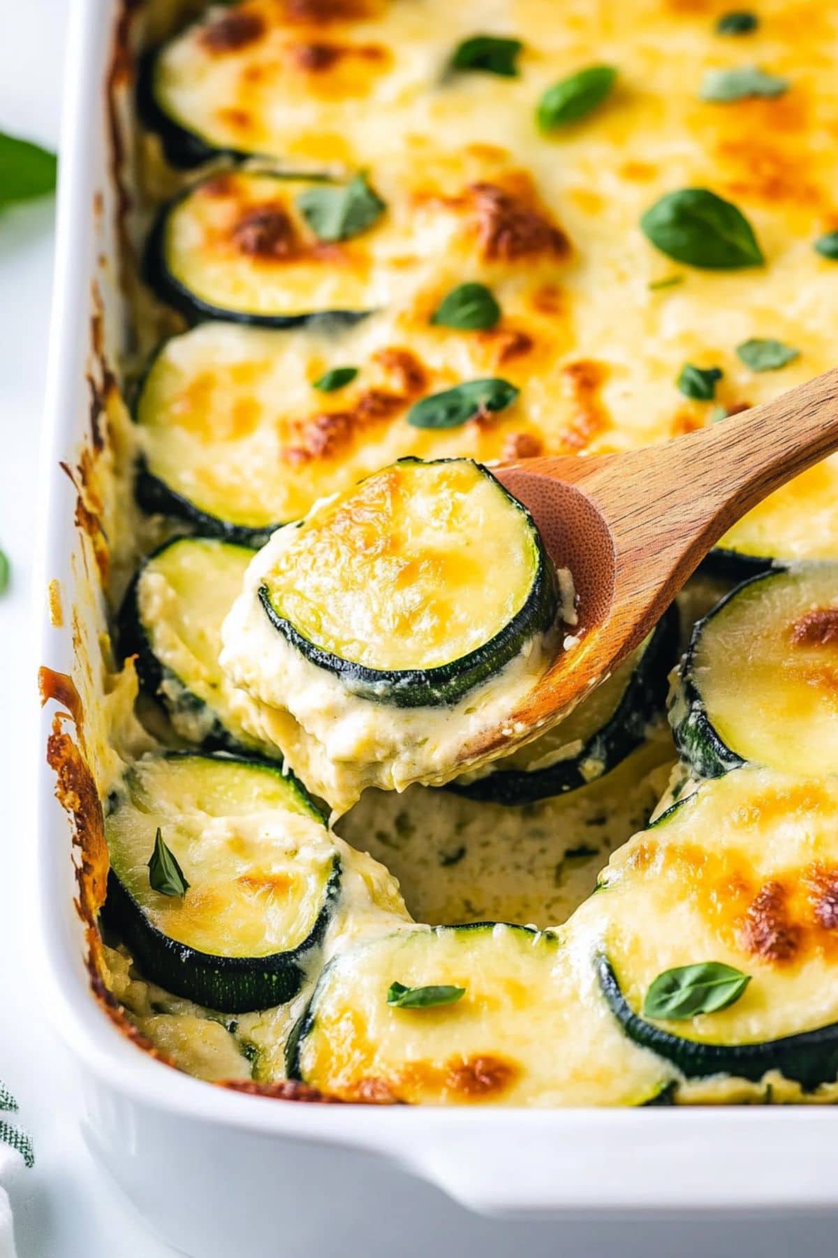 Wooden spoon lifting a scoop of cheesy scalloped zucchini from a baking dish.