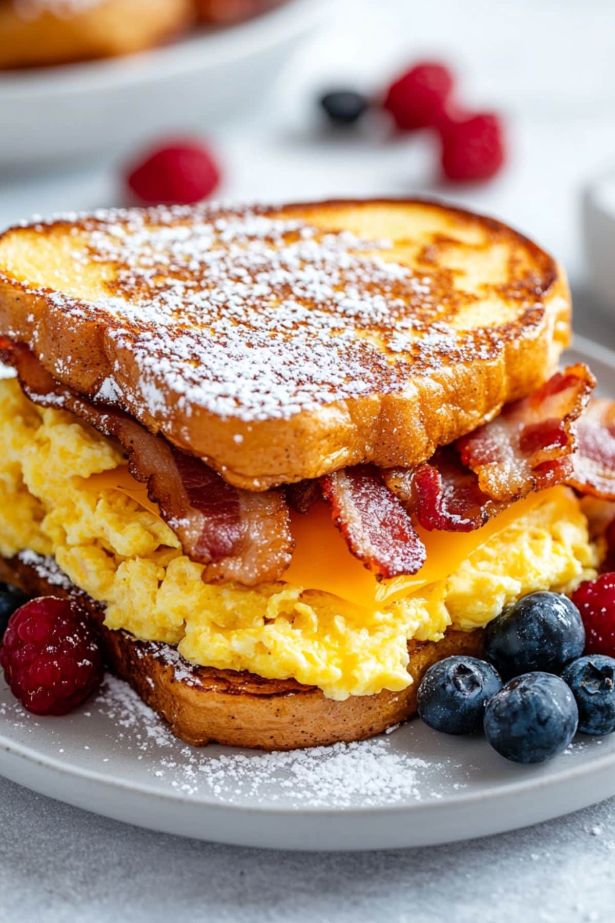 French toast breakfast sandwich with egg, bacon and cheese filling served in a white plate with berries garnish.