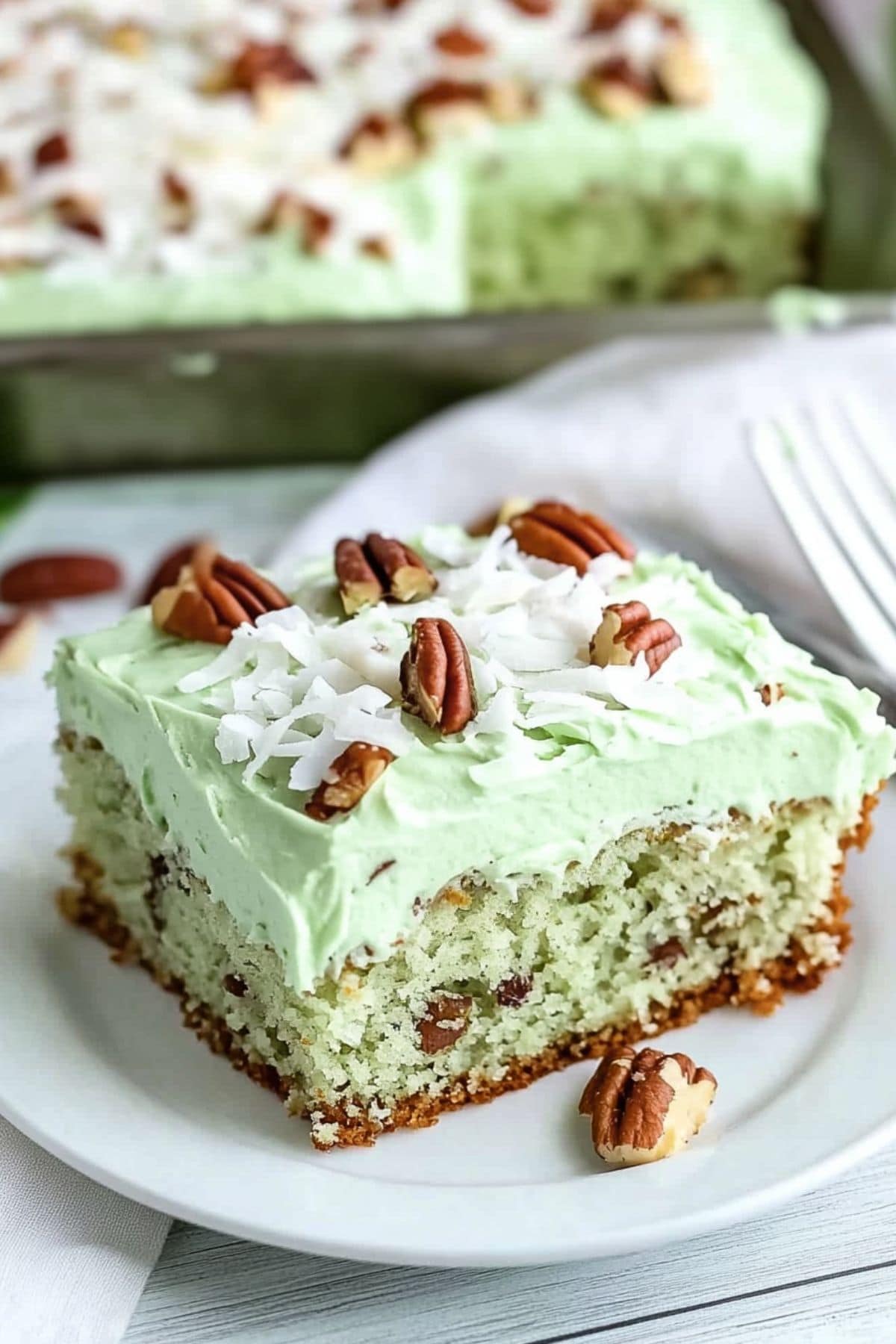 A slice of moist and fluffy watergate cake with green frosting garnished with chopped pecans and shredded coconut.