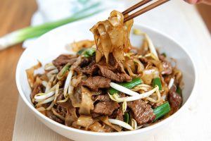 Wooden chopsticks picking noodles from a bowl serving of beef chow fun.