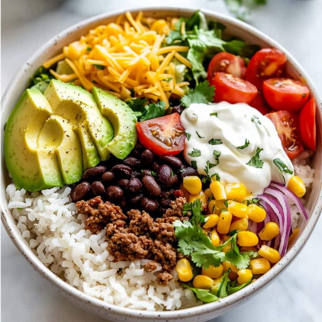 Cilantro lime rice in a white bowl topped with ground beef, black beans, corn and avocado garnished with chopped parsley and sour cream.