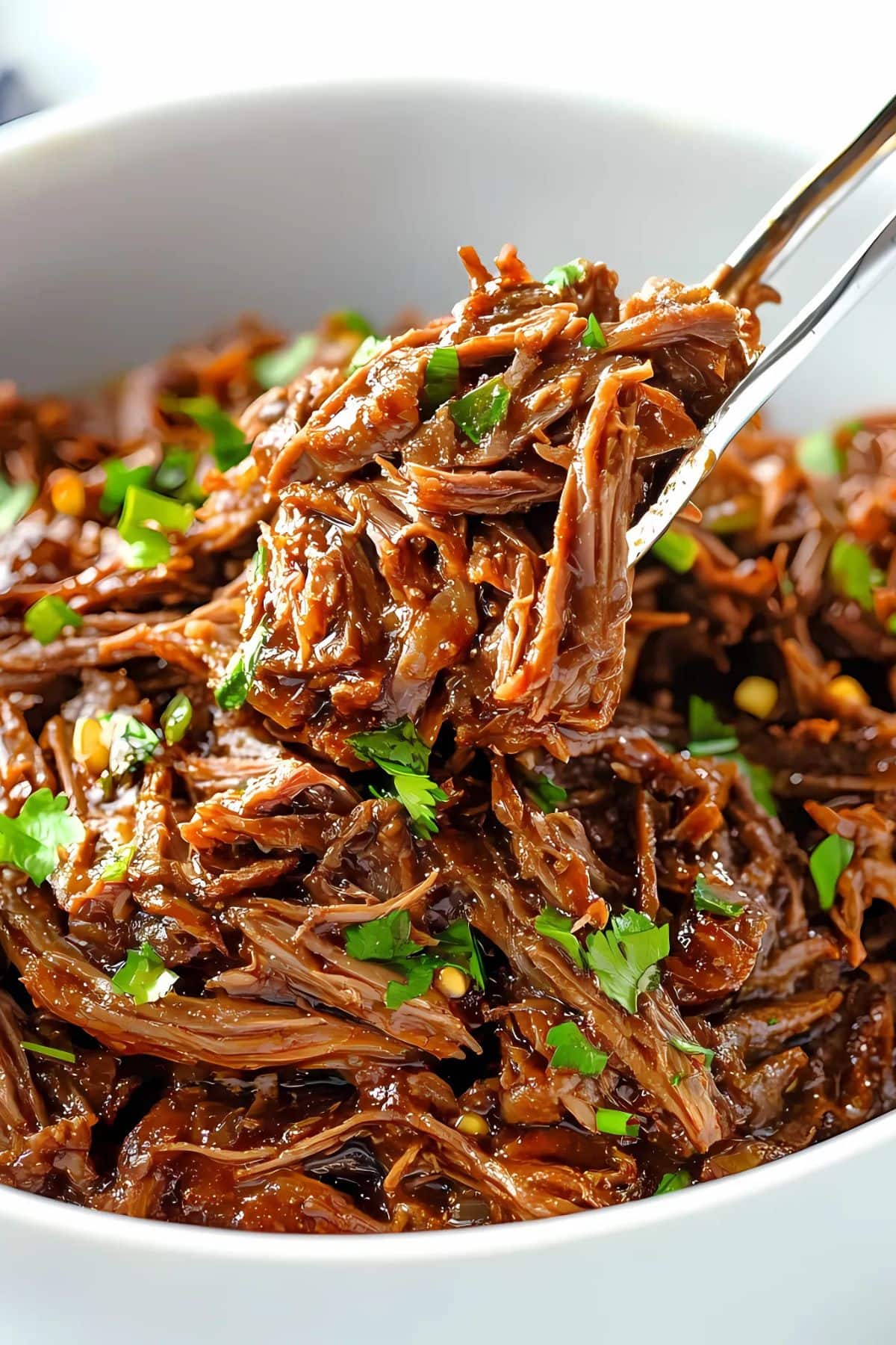 Barbacoa beef in a white bowl lifted by a metal tong.