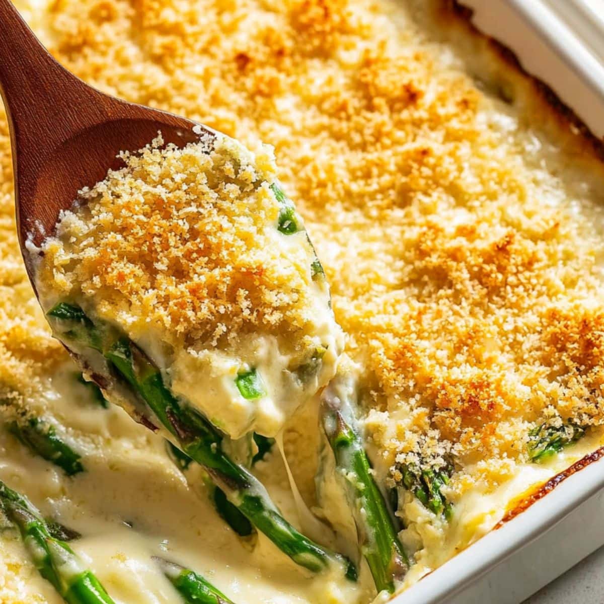 Wooden ladle scooping creamy asparagus casserole from baking dish.