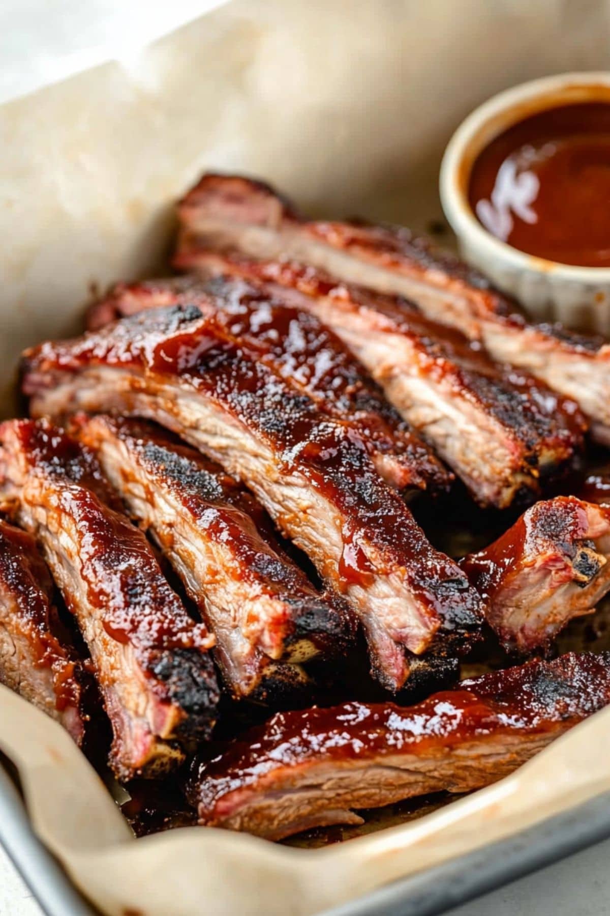 Sliced baby back pork ribs arranged in a pan with parchment paper serve with barbeque sauce. 