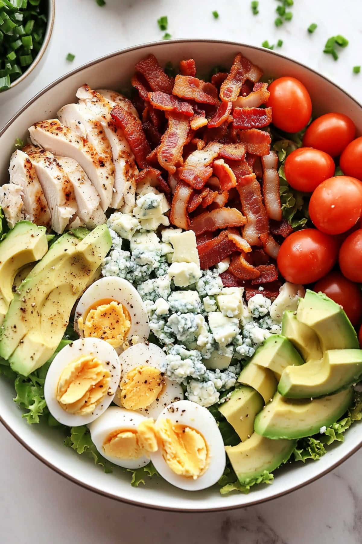 Cobb Salad featuring chicken, romaine lettuce, bacon, eggs, avocados, cherry tomatoes, blue cheese and chopped chives in a bowl on a white marble table.